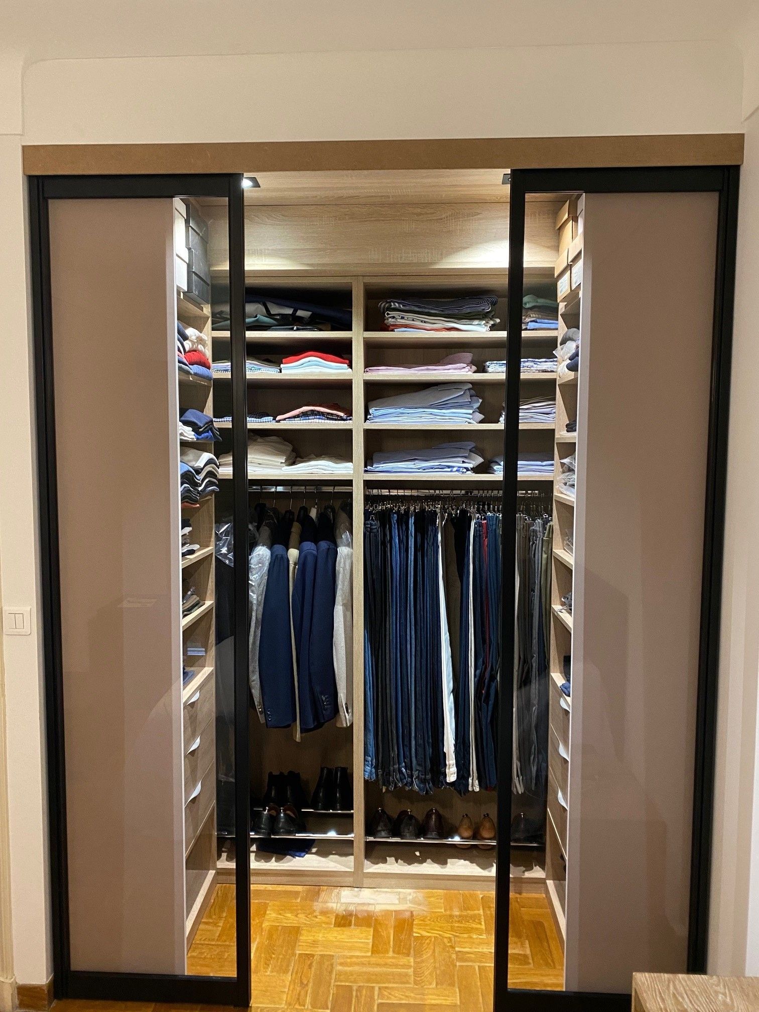 Dressing avec portes coulissantes. Murs beiges, cadres noirs, étagères en bois avec vêtements et chaussures.