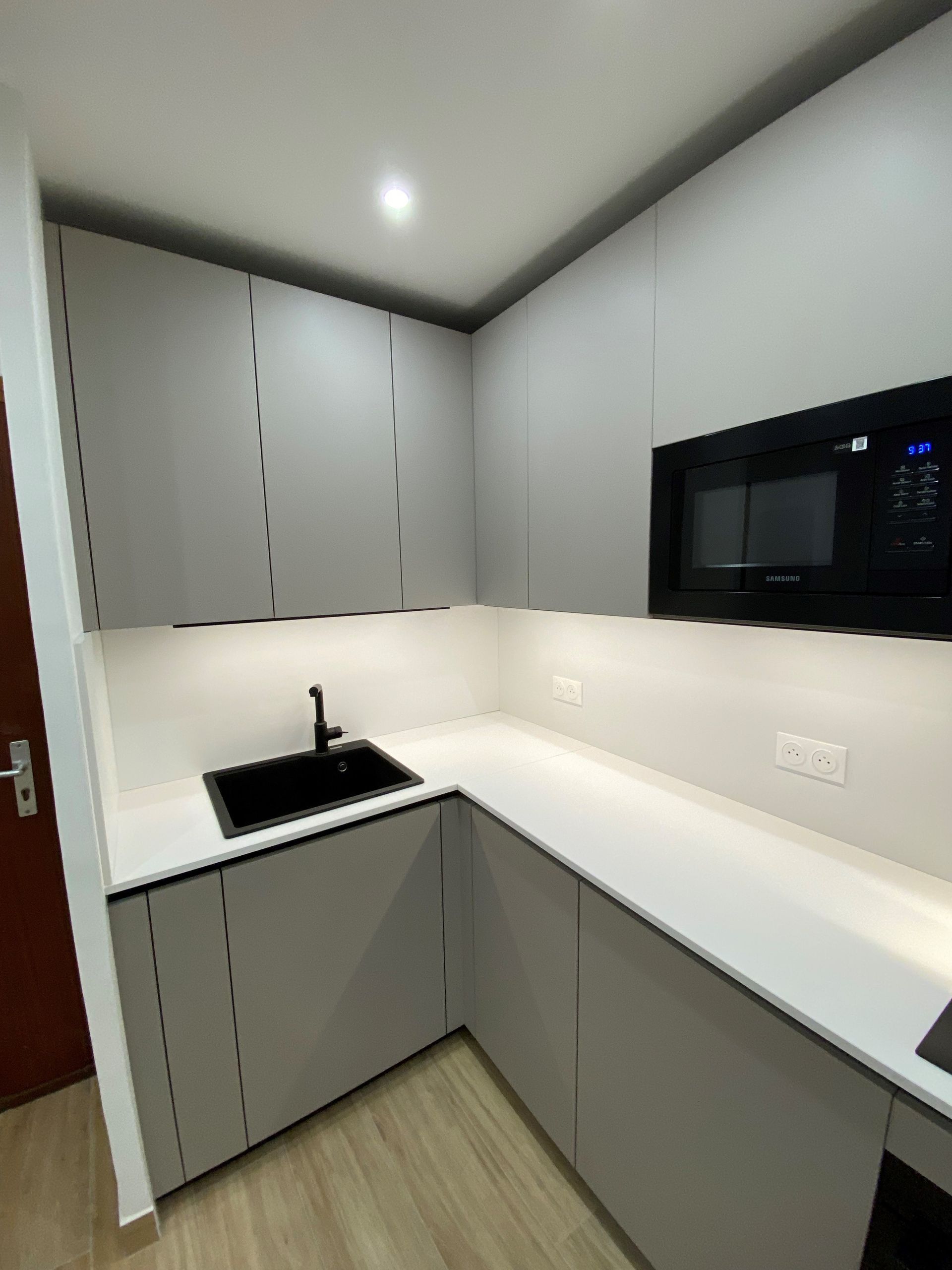 Cuisine moderne et minimaliste avec armoires grises, comptoirs blancs et évier et micro-ondes noirs.