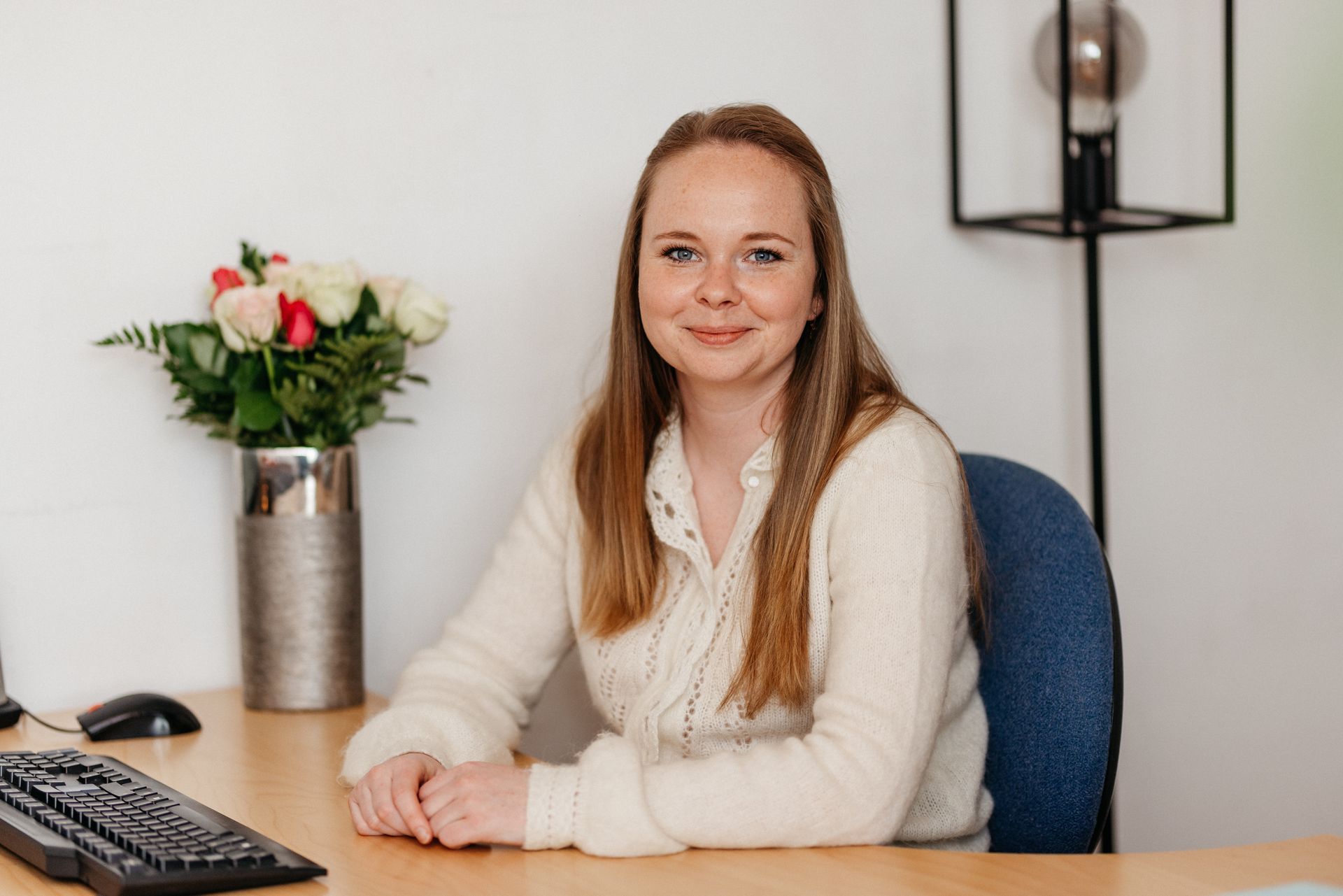 Femme aux cheveux longs dans un pull crème, assise à un bureau avec un bouquet de roses.