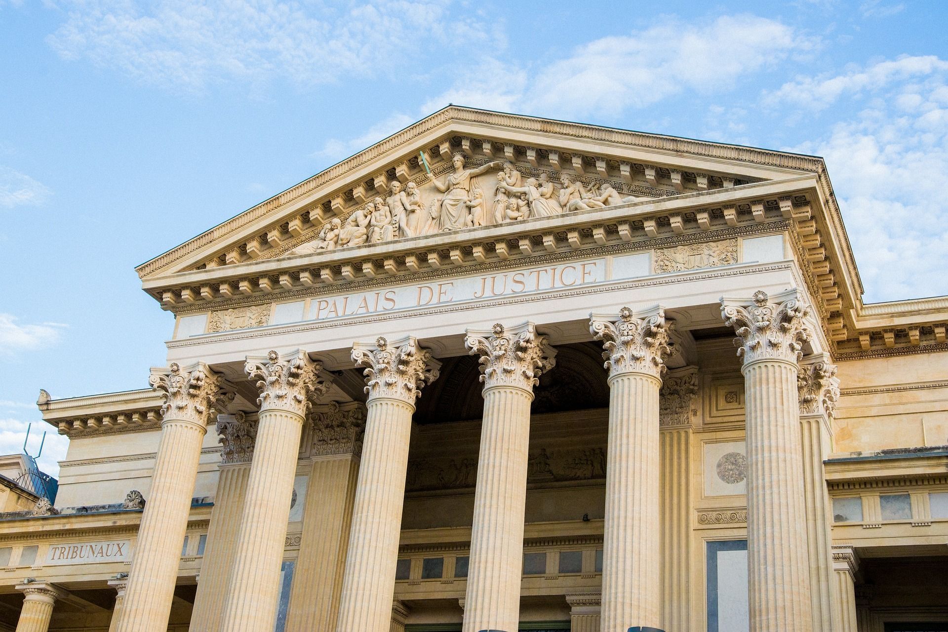 Extérieur d'un palais de justice classique avec des colonnes et un fronton sous un ciel partiellement nuageux.