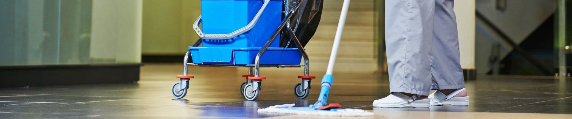 Une personne lave le sol mouillé. Un seau bleu sur un chariot à roulettes se trouve à proximité, dans un couloir.