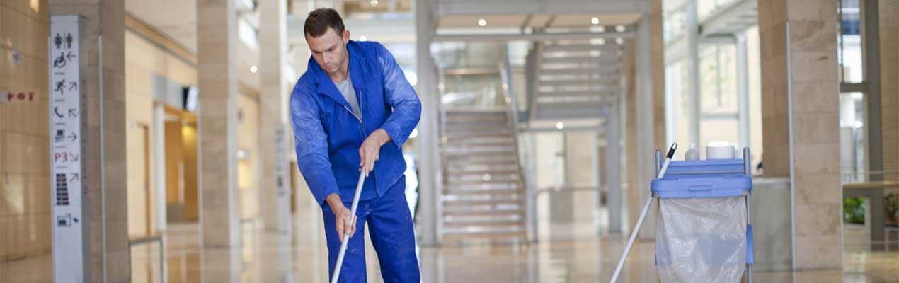 Une personne en salopette bleue nettoie un grand couloir bien éclairé avec une serpillière et un seau.