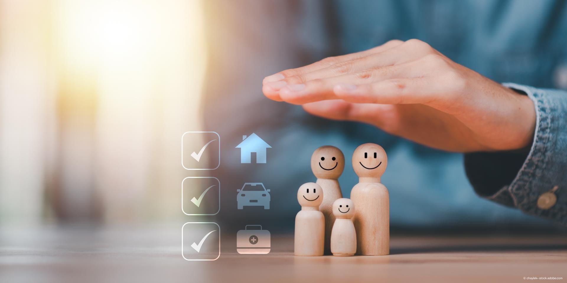 Ein Versicherungsmitarbeiter hält schützend die Hand über 4 hölzerne Personen, welche eine Familie darstellen