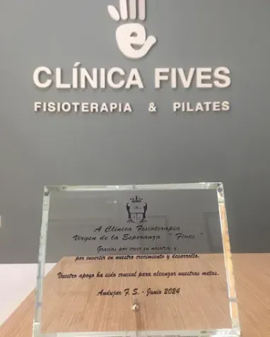 Rótulo de clínica y premio: “Clínica Fives” con “Fisioterapia & Pilates” y placa de cristal, sobre pared gris.