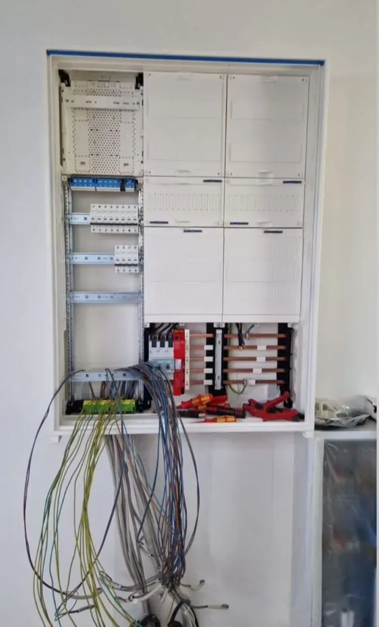 Offener Elektro-/Netzwerkverteiler mit freiliegenden Kabeln und beschrifteten Modulen, an einer Wand montiert.