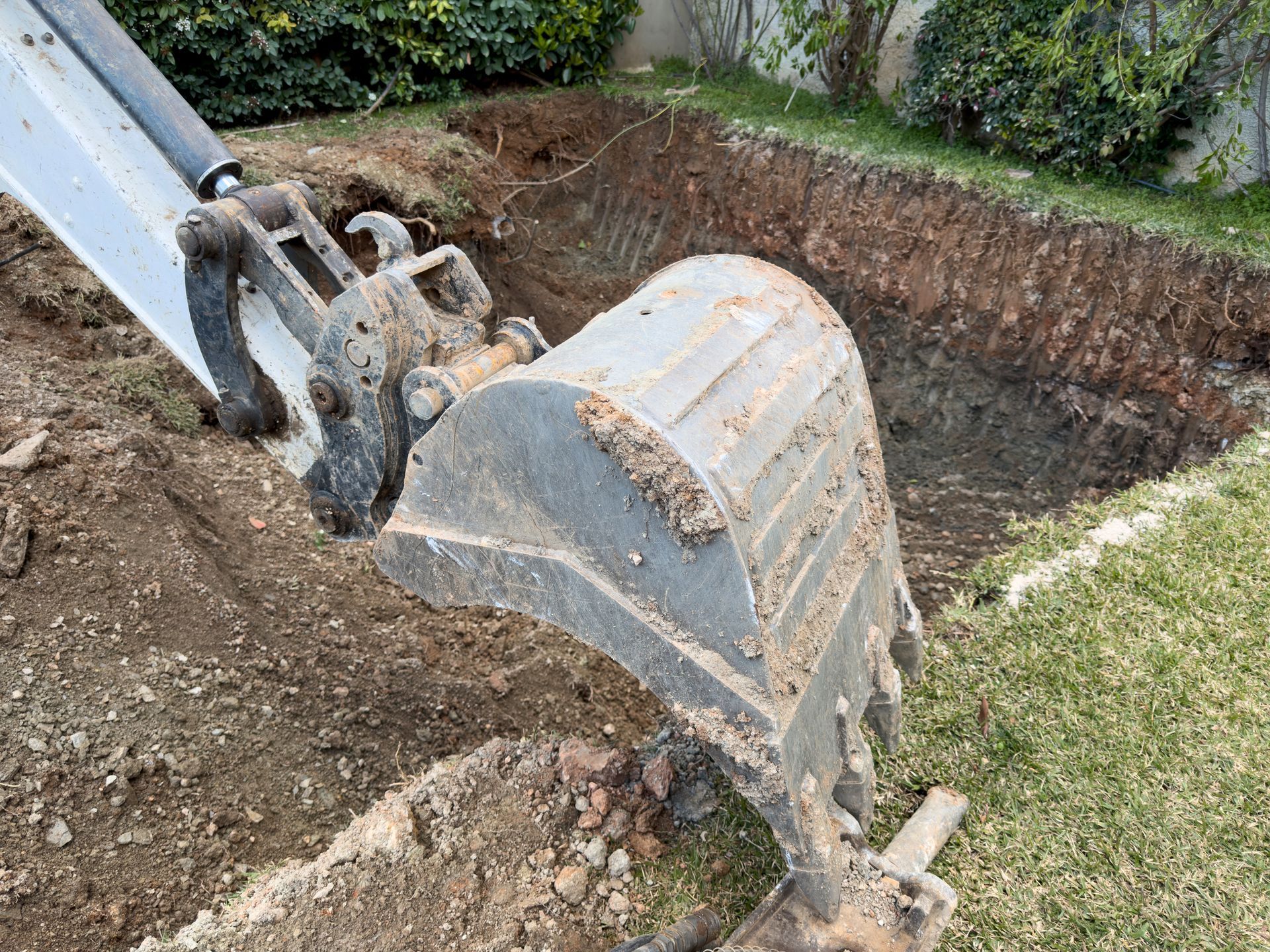 Godet d'une excavatrice creusant une tranchée rectangulaire dans une cour en terre battue, près d'une bordure recouverte d'herbe