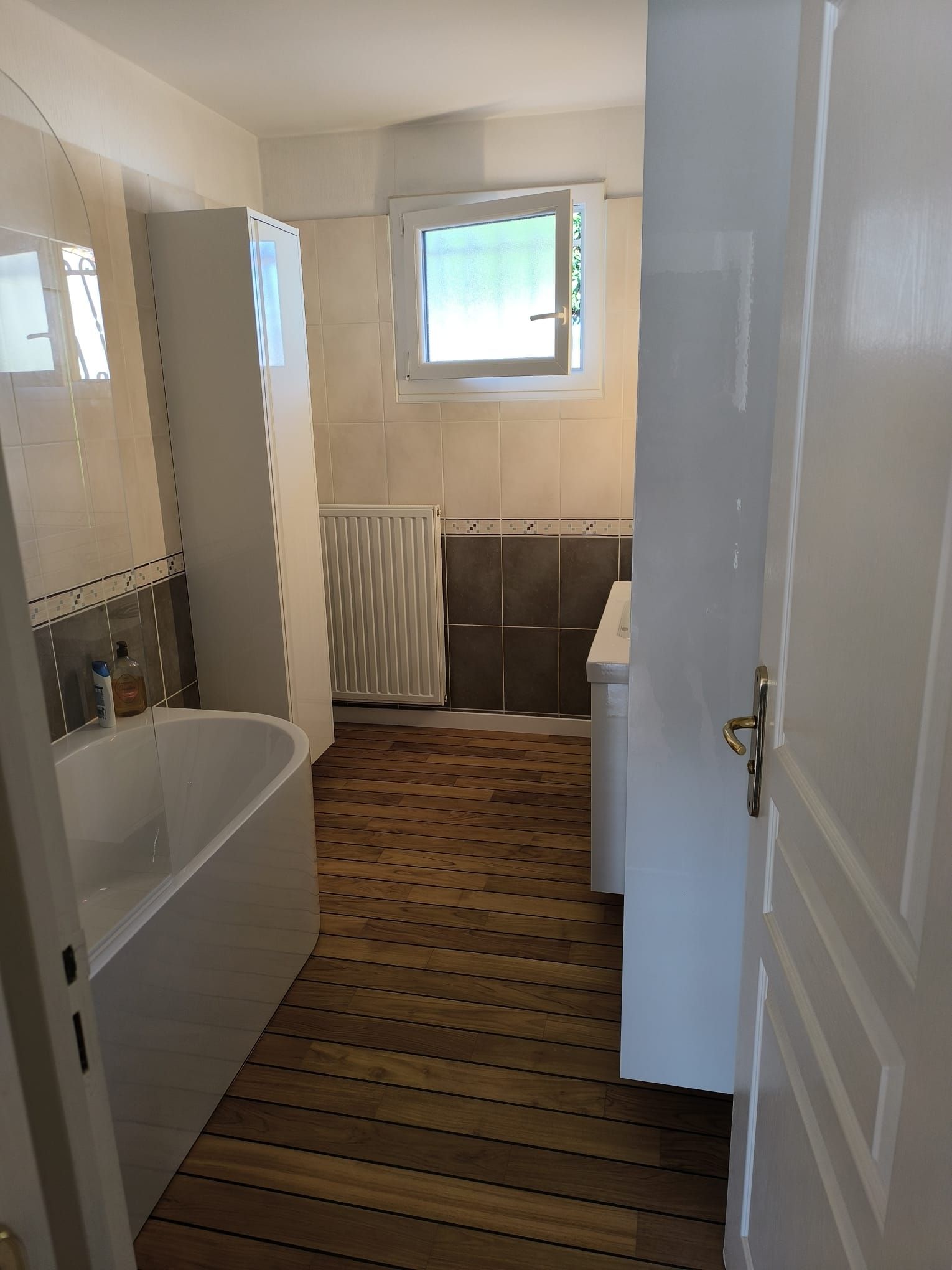 Salle de bains avec baignoire, radiateur, fenêtre et sol carrelé marron.