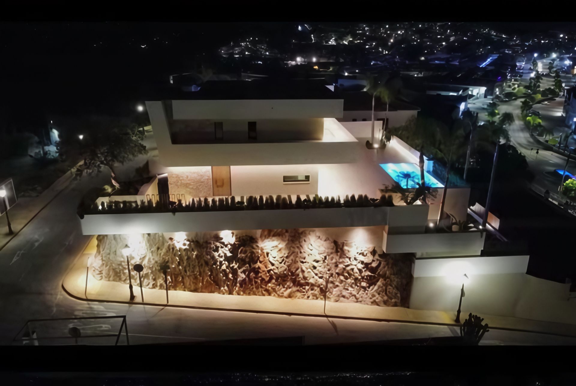 Moderna casa blanca de noche con piscina, fachada de piedra y luces de la ciudad de fondo.