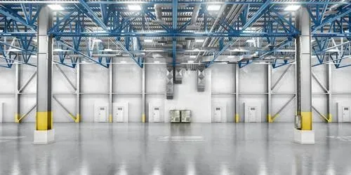 Interior vacío de un almacén industrial con vigas de acero azul y postes de seguridad amarillos bajo luces brillantes.