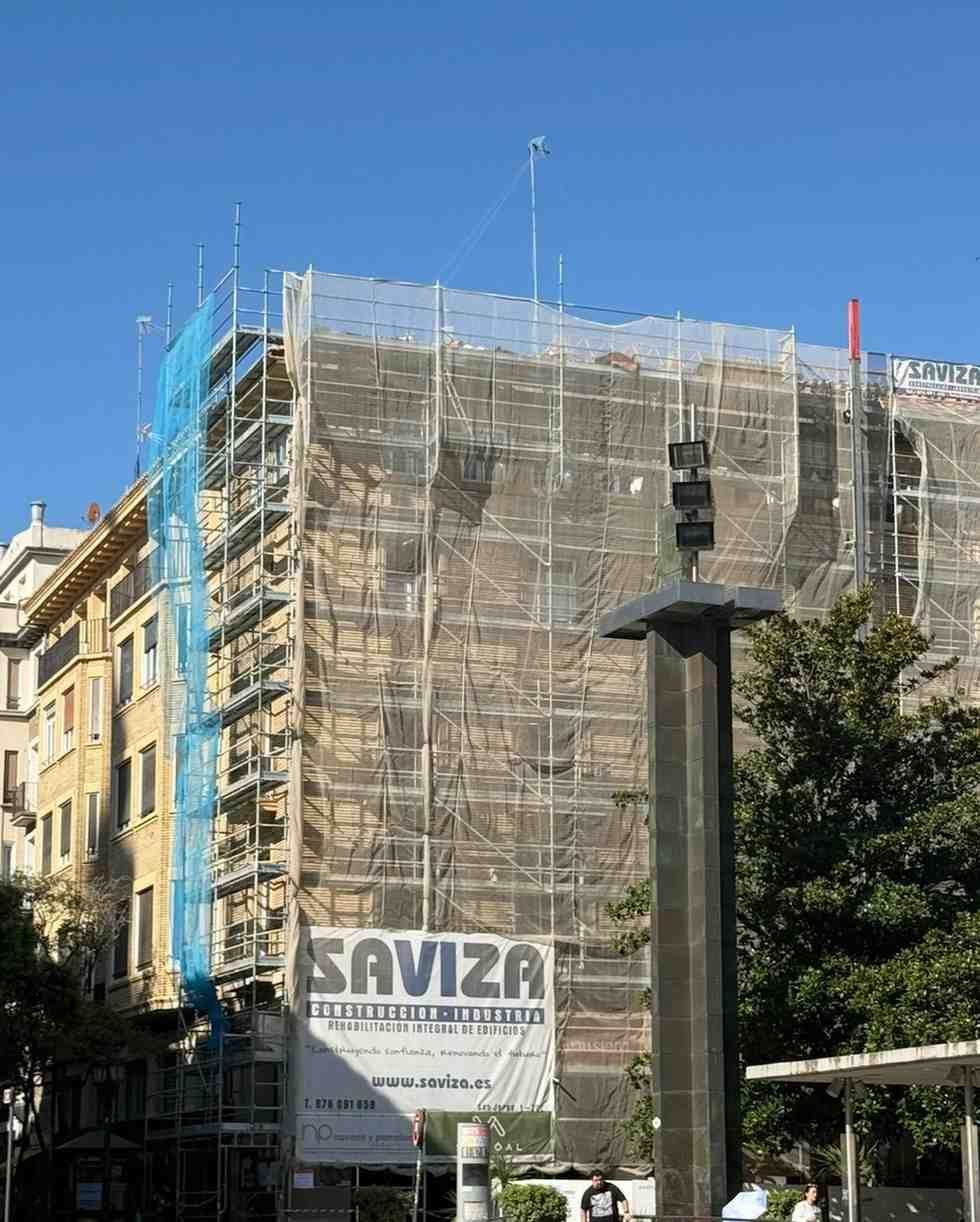 Edificio en obras, con andamios, detrás de una pancarta de “SAVIZA” en una calle soleada.