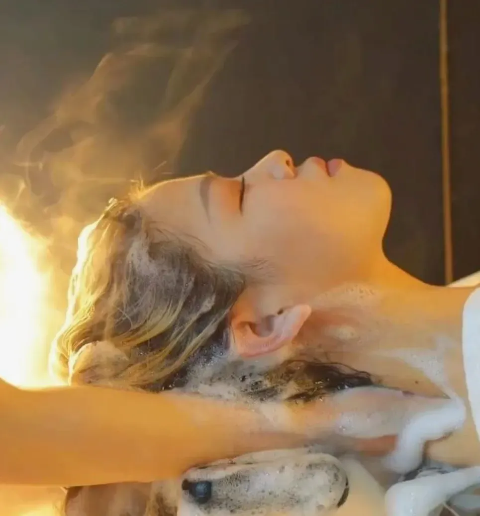 Mujer lavándose el cabello, con los ojos cerrados y con vapor elevándose en un salón de belleza.