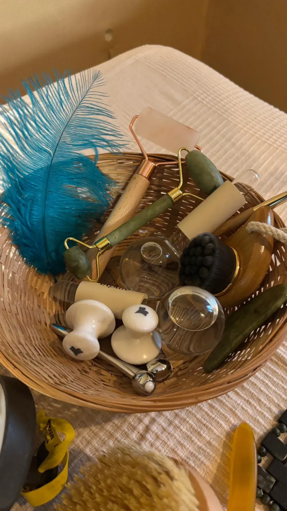 Cesta de mimbre llena de herramientas de masaje facial, incluidos rodillos y cepillos, sobre una mesa de color crema.