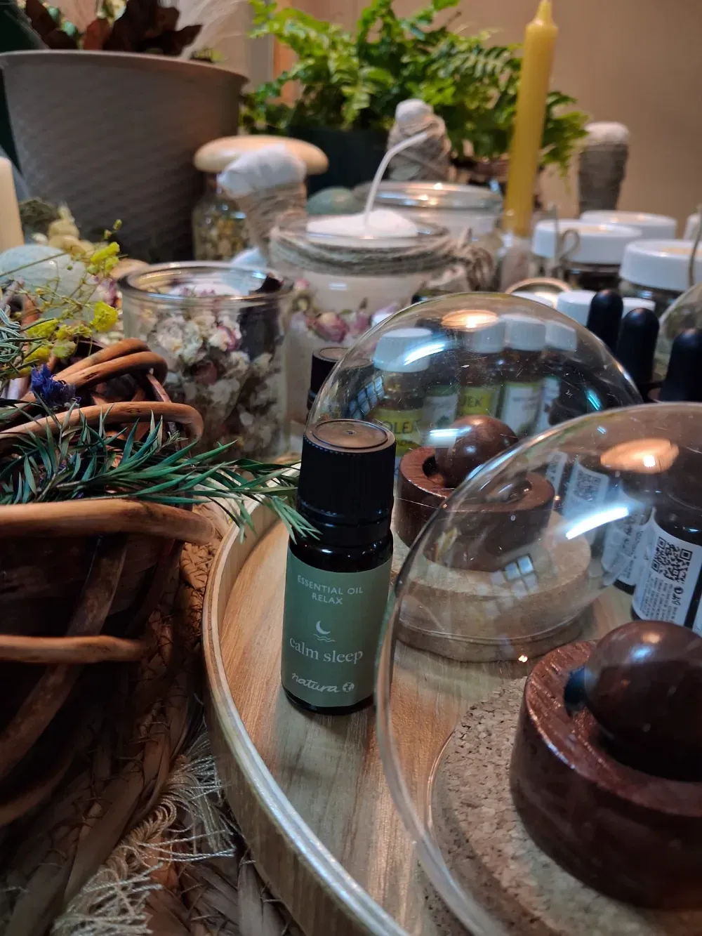 Botella de aceite esencial en una bandeja de madera rodeada de frascos, plantas y velas.