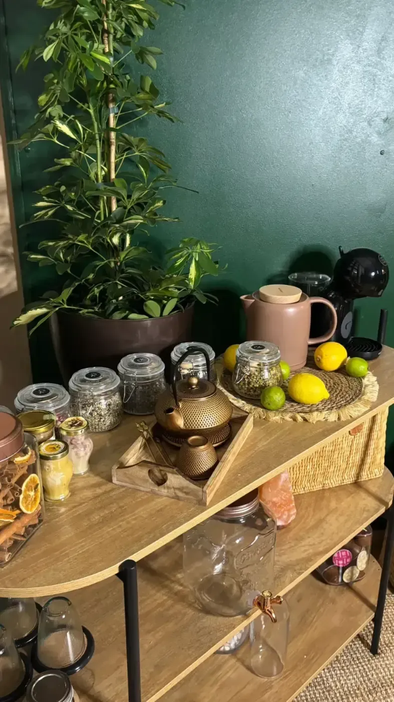 Estantería con tazas de té, tetera, frascos de hierbas, limones y una planta contra una pared verde.
