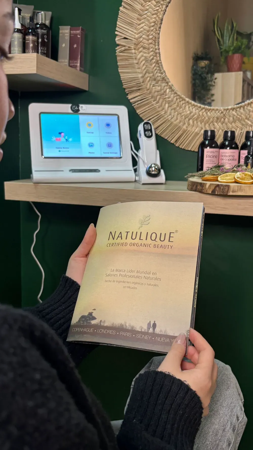 Persona con un folleto de Naturique en un spa con paredes verdes. Se ven una pantalla digital y productos.