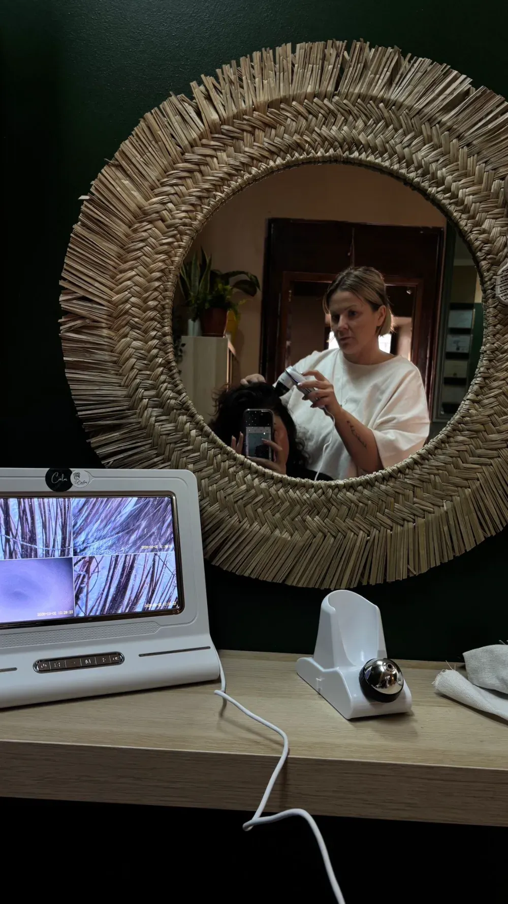 Una mujer examina su cabello bajo un espejo con un dispositivo de análisis del cuero cabelludo. Salón con pared verde.