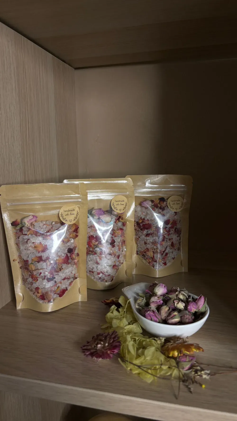 Tres bolsas de sales de baño con pétalos de flores en un estante. Un cuenco con flores secas y pétalos está enfrente.
