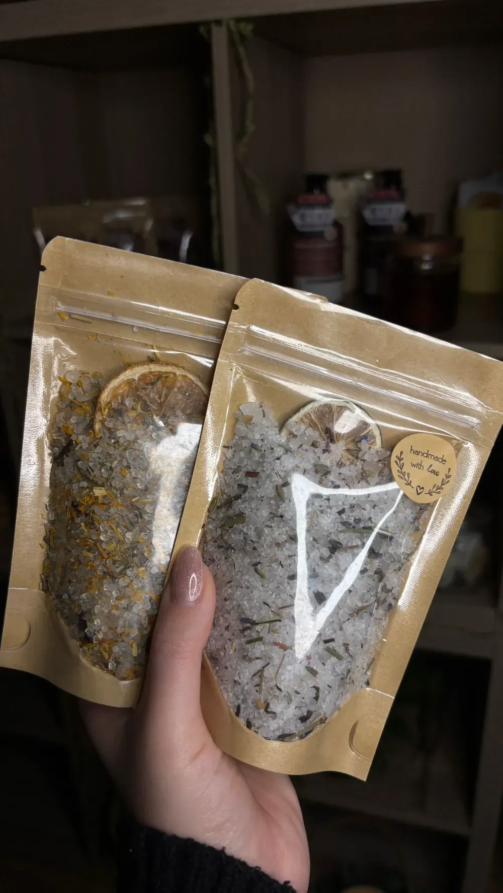 Dos bolsas marrones con sales de baño, sostenidas por una mano; estante al fondo.