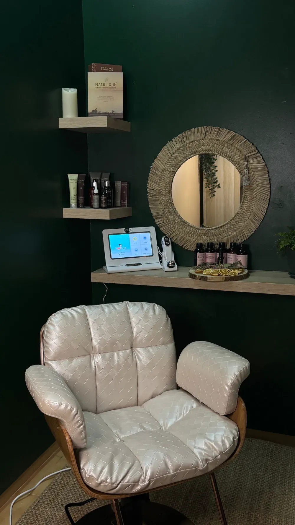 Acogedor rincón de spa con silla blanca, estantes con productos, espejo redondo sobre pared verde.