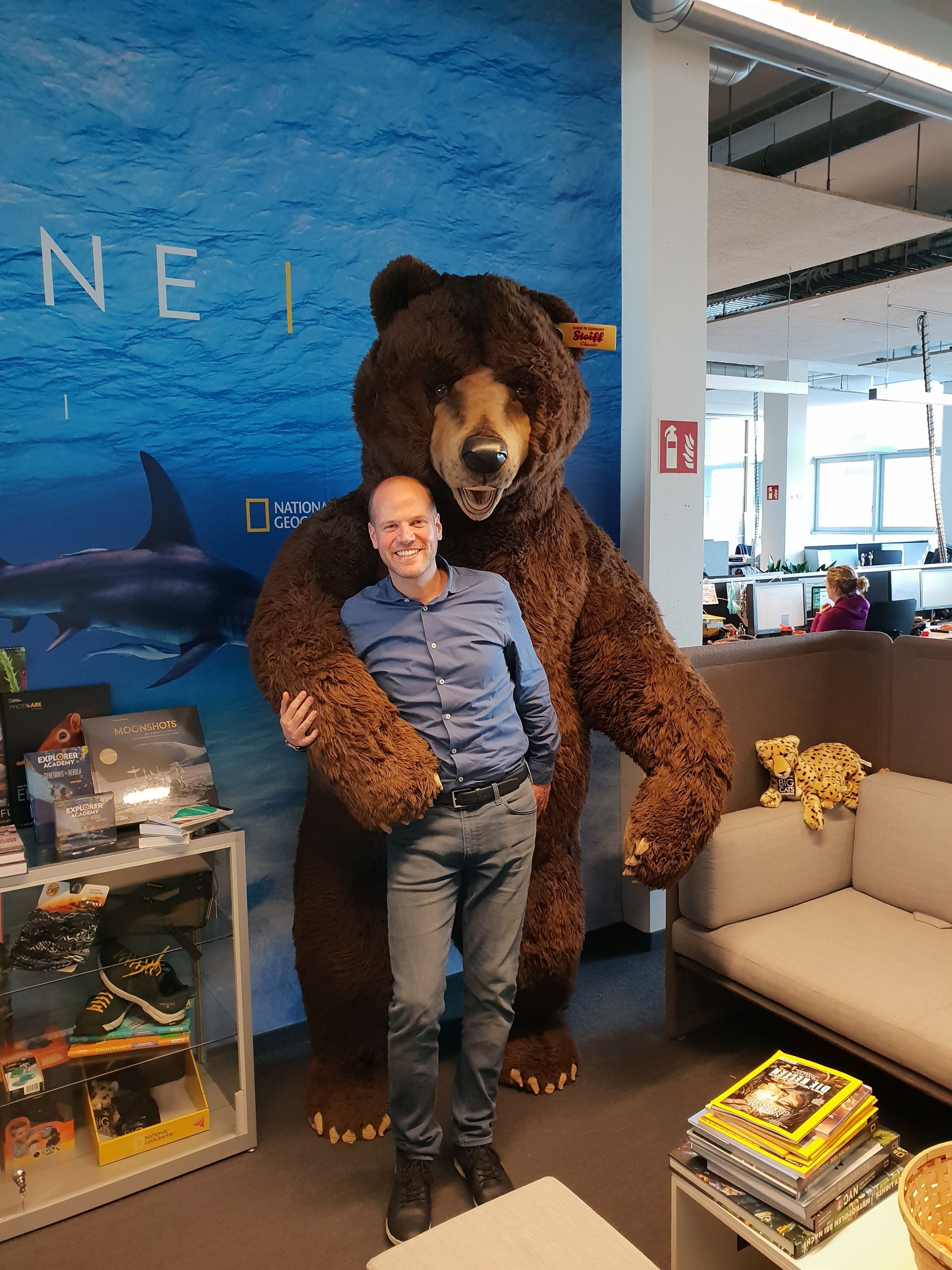Mann umarmt großen ausgestopften Braunbären in Büroumgebung, blauer Hintergrund und Zeitschriften auf dem Boden.
