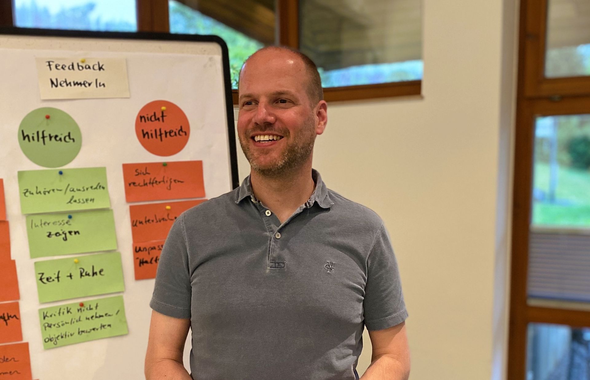 Mann mit Glatze lächelt vor einem Whiteboard mit bunten Haftnotizen in einem Konferenzraum.