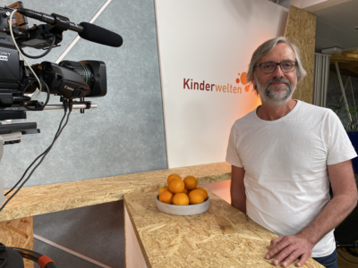 Mann vor einem Set für „Kinderwellen“, mit Kamera, Orangen und schlichtem Hintergrund.
