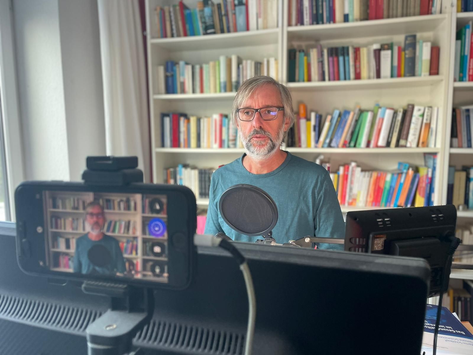 Mann mit grauem Haar und Brille nimmt vor einem Bücherregal ein Video auf.