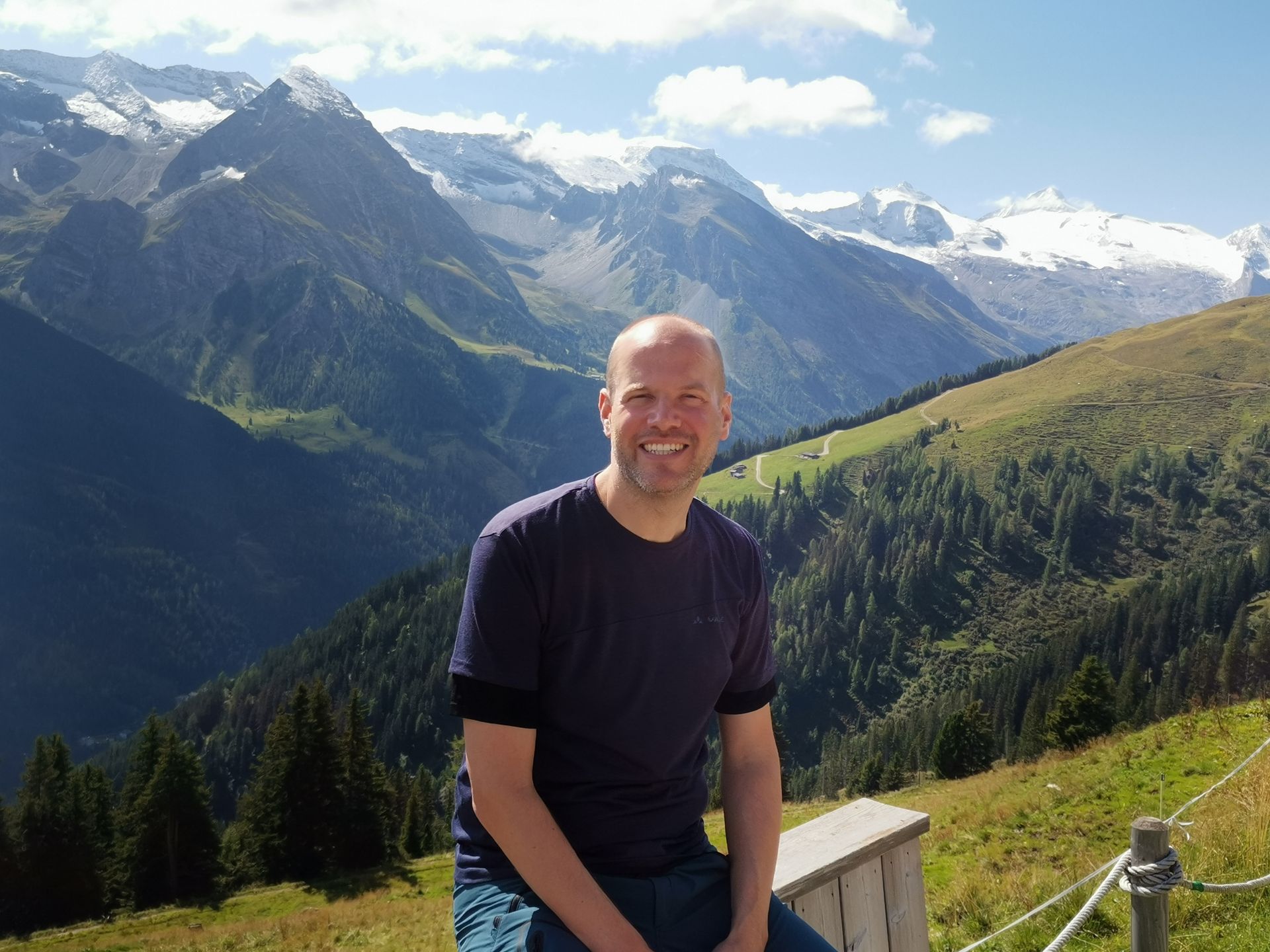 Mann lächelt in den Bergen. Grüner Hang, schneebedeckte Gipfel. Blaues Hemd, heller Tag.