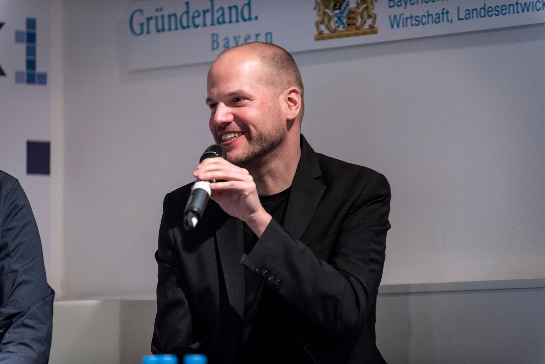 Mann mit heller Haut lächelt, hält ein Mikrofon, trägt einen schwarzen Blazer und spricht auf einer Konferenz.