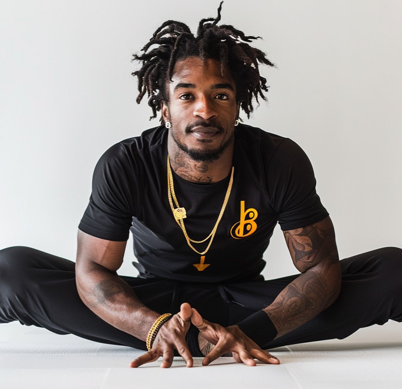 Mann in Yoga-Pose, schwarze Kleidung, Goldketten, Tattoos, Dreadlocks, neutraler Hintergrund.