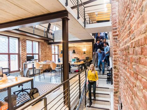 Menschen in einem Büro mit unverputzten Ziegelwänden, die Treppen hinauf- und hinuntergehen; eine Frau in Gelb.
