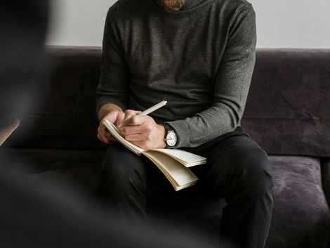 Mann im grauen Pullover, schreibt in ein Notizbuch auf einer Couch, möglicherweise in Therapie.