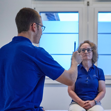 Eine medizinische Fachkraft in blauer Uniform hilft einer Person in grauer Jogginghose bei einer Beindehnübung auf einer Behandlungsliege.