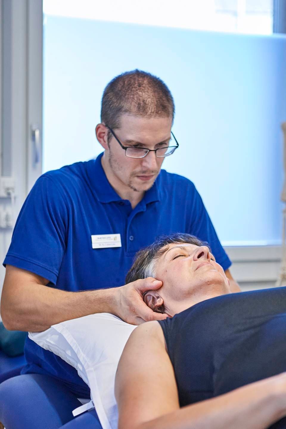 Ein Therapeut in einem blauen Poloshirt führt eine manuelle Nackenkorrektur an einem Patienten durch, der auf einer Behandlungsliege liegt.
