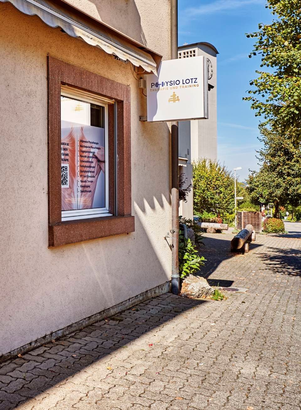 Ein hellbraunes Gebäude mit einem weissen Hängeschild mit goldenem Emblem, neben einem Schaufenster an einem Kopfsteinpflasterweg.