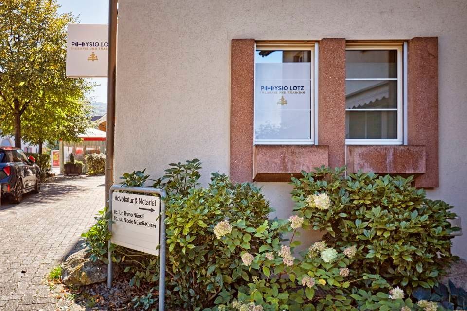 Eine helle Gebäudefassade mit einem „Pilates Lift“-Schild an einem Fenster und einem Pfeil, der auf einen Weg weist.