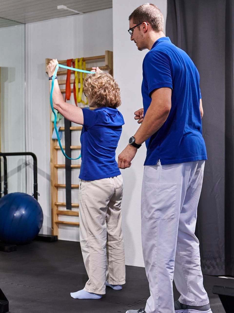 Ein Profi in einem blauen Hemd unterstützt eine Person in einem blauen Hemd bei der Physiotherapie mit einem Widerstandsband.