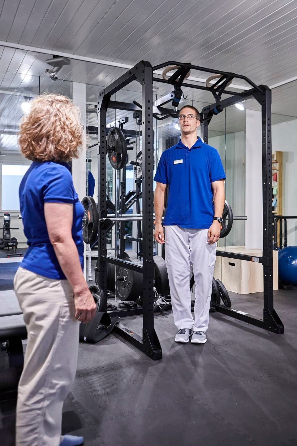 Ein Physiotherapeut beobachtet einen Patienten, der in aufrechter, neutraler Position in einem Power Rack im Fitnessstudio steht.