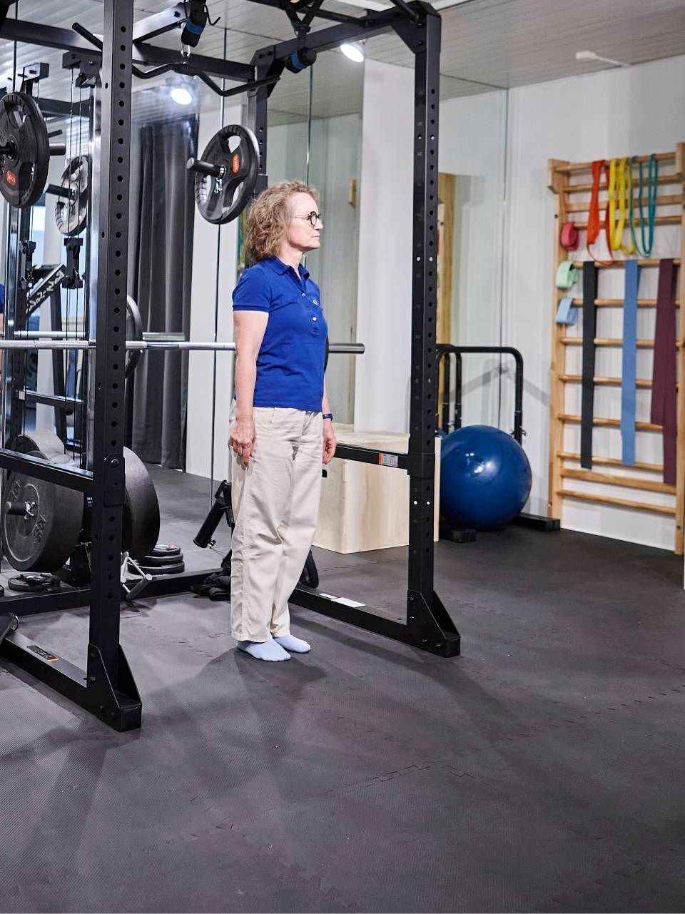 Eine Person steht in einem Power Rack eines Fitnessstudios neben einem an der Wand befestigten Ständer mit Fitnessbändern und einem blauen Gymnastikball.