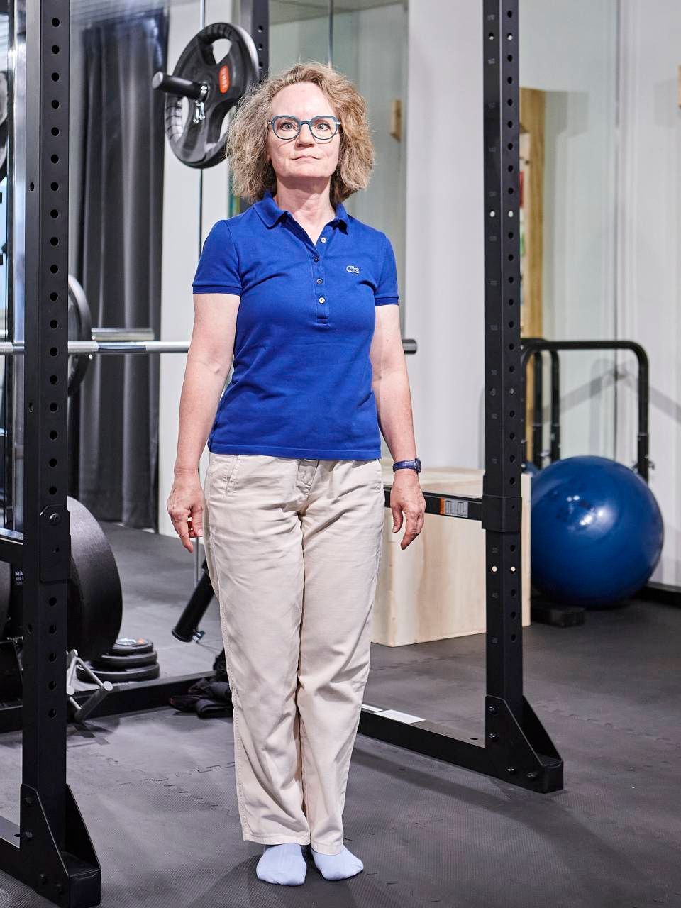 Eine Person in einem blauen Poloshirt und einer hellen Hose steht in einem Fitnessstudio zwischen den Ständern eines Kniebeugenständers.