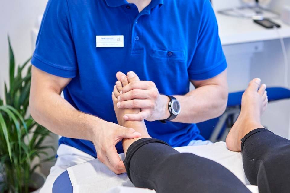 Ein Physiotherapeut in einem blauen Hemd untersucht den Fuss eines Patienten in einer klinischen Umgebung.