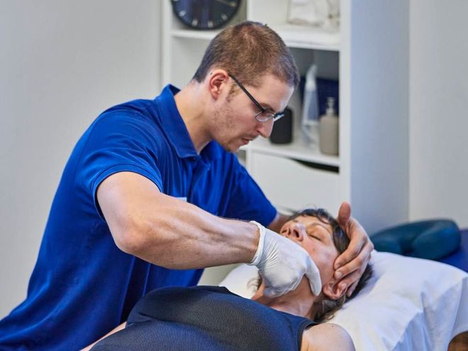 Eine medizinische Fachkraft in einem blauen Hemd führt in einer klinischen Umgebung eine intraorale manuelle Therapie am Kiefer eines Patienten durch.