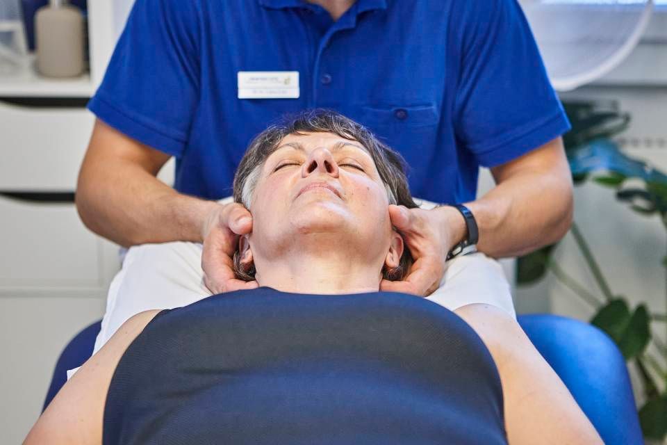 Ein Therapeut in einem blauen Poloshirt führt in einer Klinik eine Nackenkorrektur an einer auf dem Rücken liegenden Person durch.