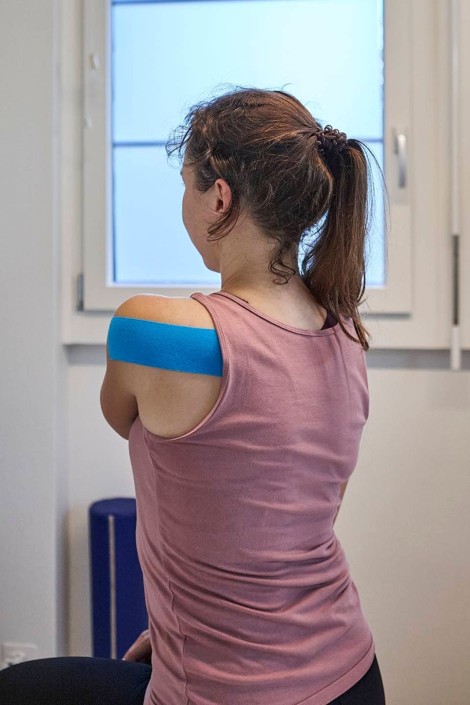 Eine von hinten gesehene Person trägt ein rosa Tanktop mit einem blauen Kinesiologie-Tape-Streifen über der Schulter.