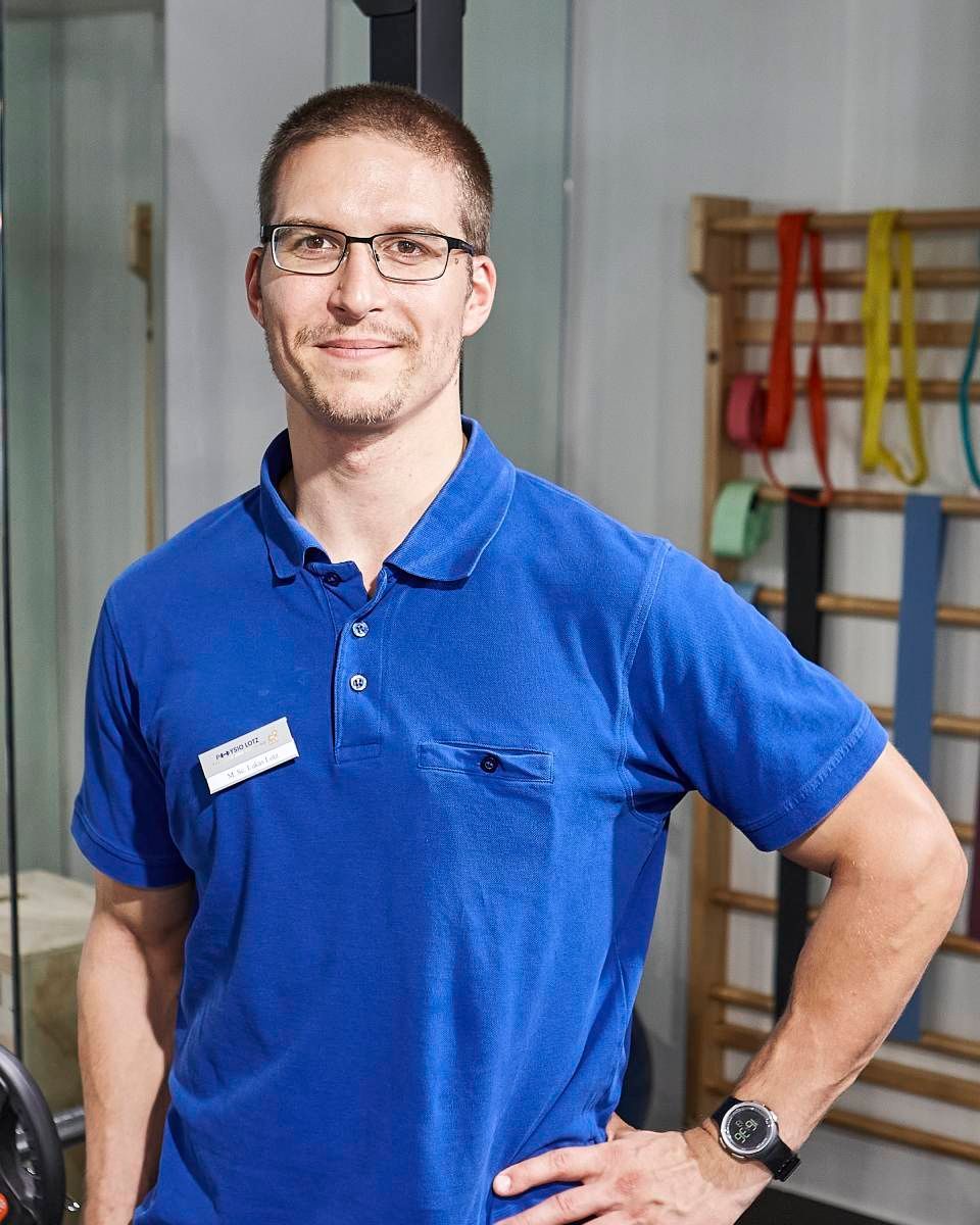 Eine Person in einem blauen Poloshirt und mit Brille steht in einem Fitnessstudio; im Hintergrund sind Widerstandsbänder zu sehen.
