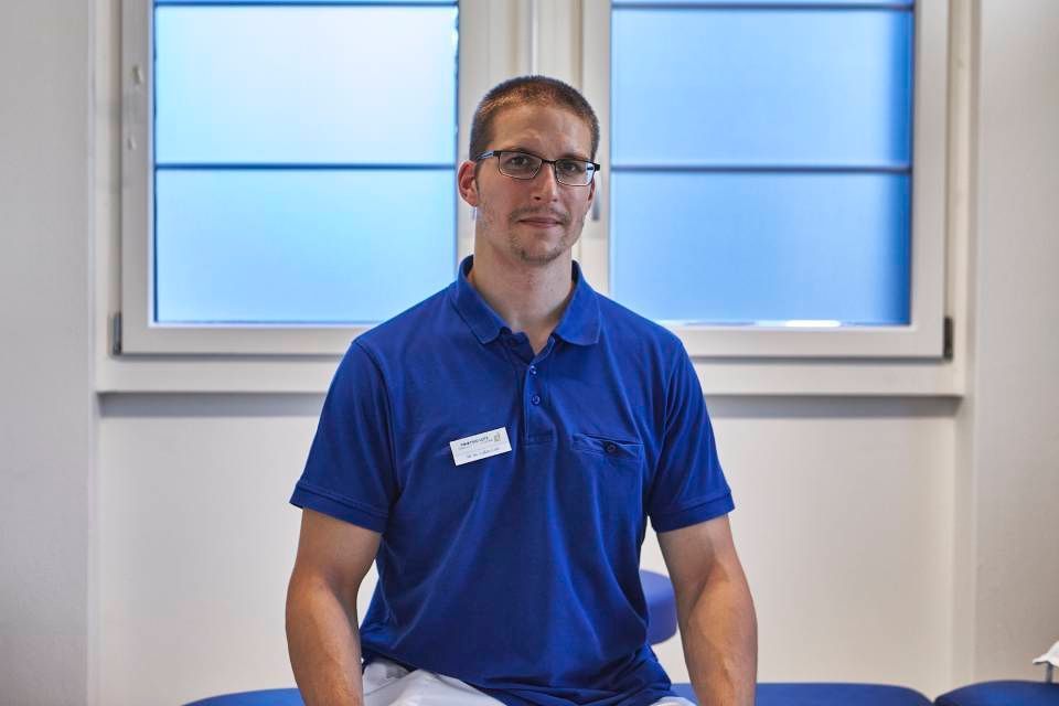 Eine Person in einem blauen Poloshirt und mit Brille sitzt in einem Büro; im Hintergrund ist ein Fenster zu sehen.