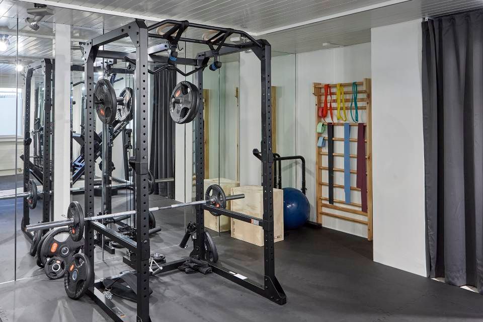 Ein Heim-Fitnessstudio mit schwarzem Kniebeugenständer, Langhantel, Gewichtsscheiben, einer hölzernen Wandleiter mit Widerstandsbändern und einem Gymnastikball.