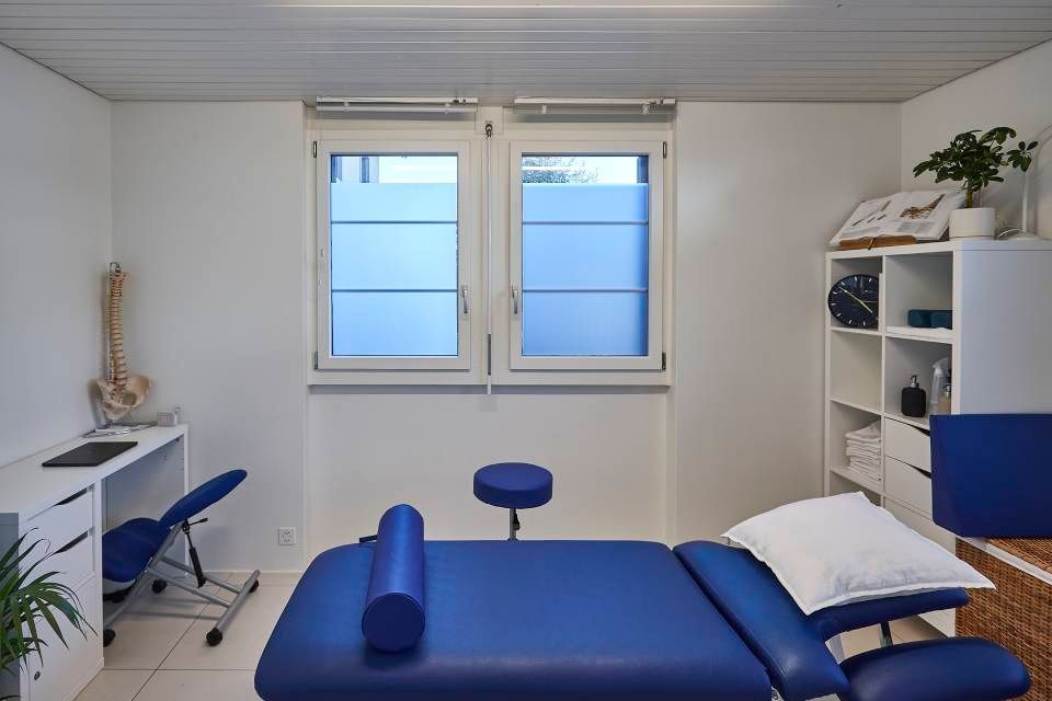Ein sauberes, weisses Arzt- oder Therapiezimmer mit einer blauen Behandlungsliege, einem Schreibtisch mit Stuhl und einem weissen Ablageregal.