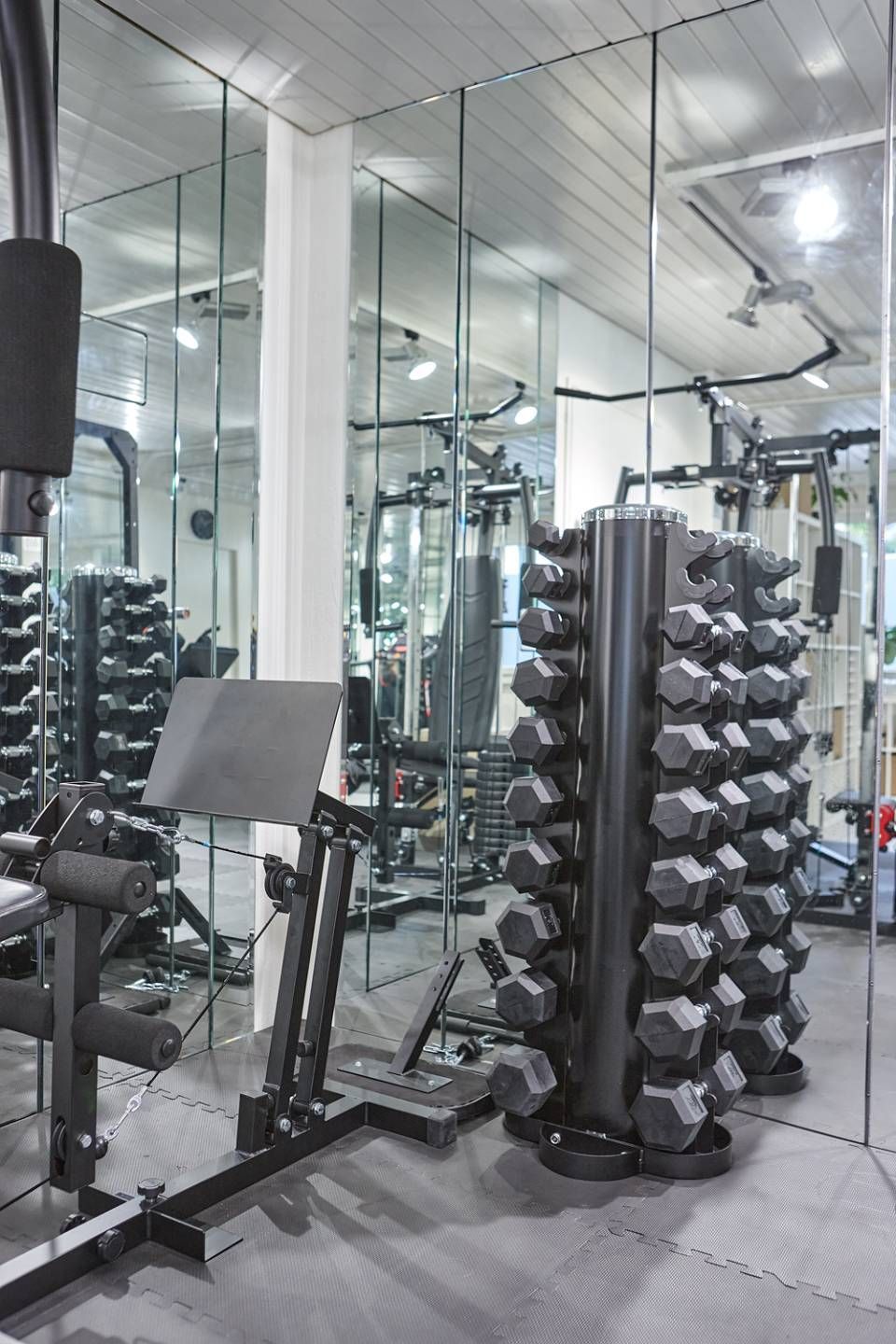 In einem Fitnessstudio steht ein Gestell mit sechseckigen Kurzhanteln, deren Spiegelbilder sich in einer Wand aus Spiegeln spiegeln, neben einer Hantelbank und einer Kabelzugmaschine.