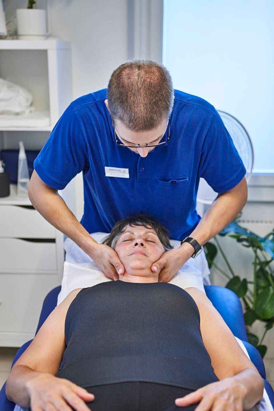 Eine medizinische Fachkraft in einem blauen Hemd führt eine Nackenkorrektur an einem Patienten durch, der auf einer Behandlungsliege liegt.